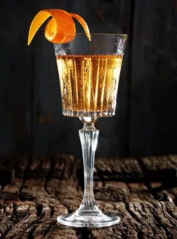 Gylden cocktail i h&oslash;jt krystalglas med appelsinskalgarnish p&aring; rustik tr&aelig;bund foran m&oslash;rk baggrund.