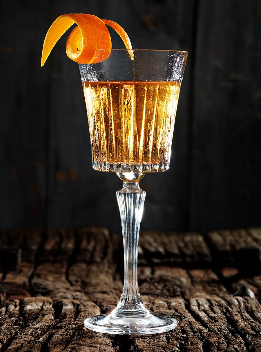 Gylden cocktail i h&oslash;jt krystalglas med appelsinskalgarnish p&aring; rustik tr&aelig;bund foran m&oslash;rk baggrund.