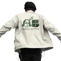 Person if&oslash;rt beige jakke med KUKU Iranian Kitchen logo trykt p&aring; ryggen.