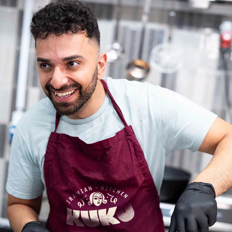 KUKU Iranian Kitchen &ndash; medarbejder i k&oslash;kkenet if&oslash;rt forkl&aelig;de med logo designet af Comugraph.