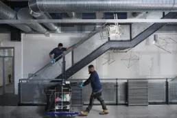 To medarbejdere fra Rengoering.com udfører professionel rengøring i en moderne industribygning med ståltrapper og ventilationsrør. Billedet viser virksomhedens fokus på kvalitet og teamwork.