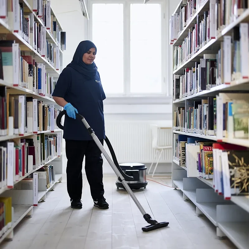 Medarbejder fra Rengoering.com st&oslash;vsuger mellem bogreoler i et lyst bibliotek. Billedet illustrerer professionel reng&oslash;ring i offentlige milj&oslash;er med fokus p&aring; orden og kvalitet.