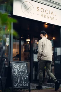 Person går ind på Social Brew café under facadeskiltet med logo designet af Comugraph – moderne cafébranding med varm og indbydende atmosfære.