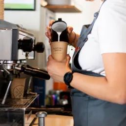 Barista hælder mælk i en to-go kop med Social Brew logo designet af Comugraph, ved espressomaskine i caféens barområde.