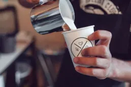 Barista hælder mælk i en kaffekop med Social Brew-logo, set tæt på i caféens varme og autentiske atmosfære.