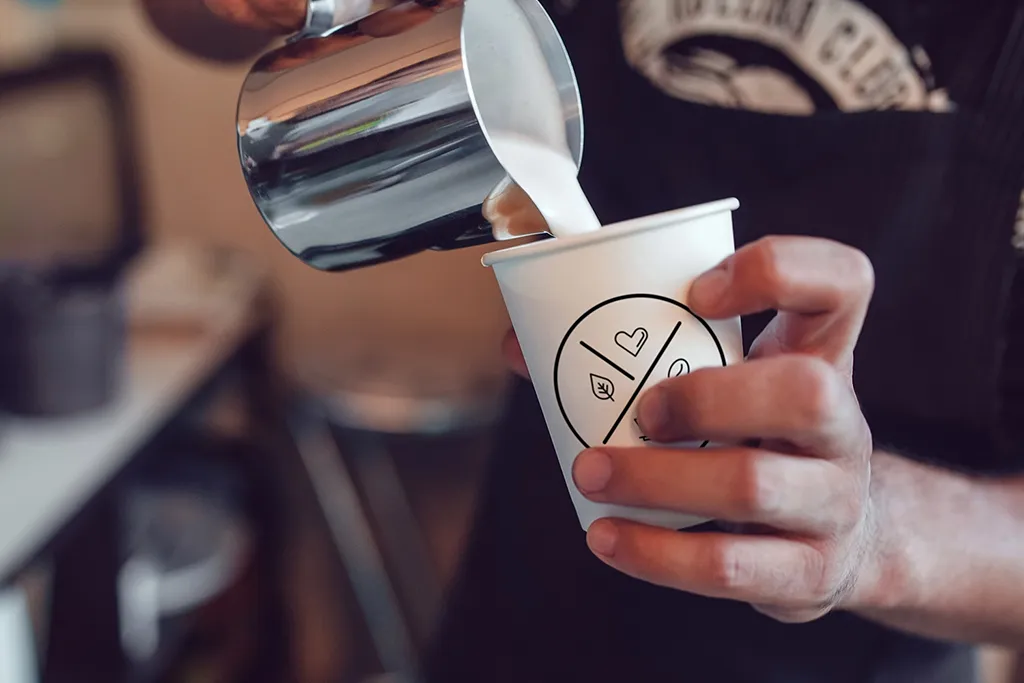 Barista hælder mælk i en kaffekop med Social Brew-logo, set tæt på i caféens varme og autentiske atmosfære.
